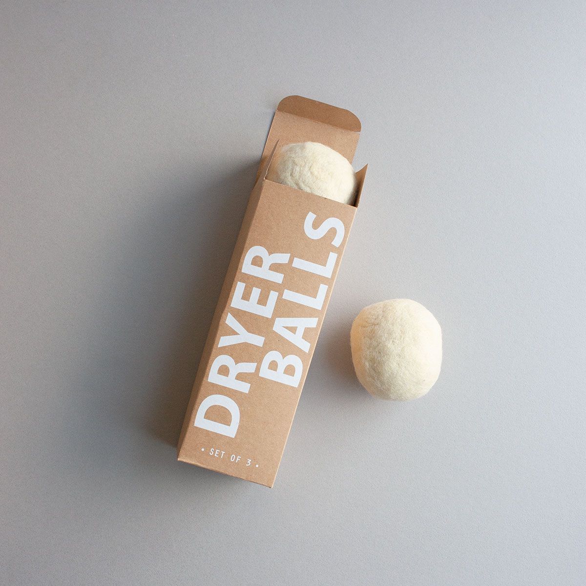 Kraft board box reads in white all-caps: Dryer Balls | Set of 3 laying on a gray background. One white felted wool ball lays outside of the box and another is peaking out of the box.