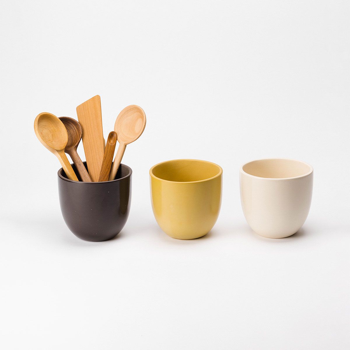 Three ceramic utensil crocks sit in a row upon a white backdrop, left to right: Woods (brown), Pollen (yellow), and Natural (cream). The brown utensil crock on the left holds various wooden utensils.