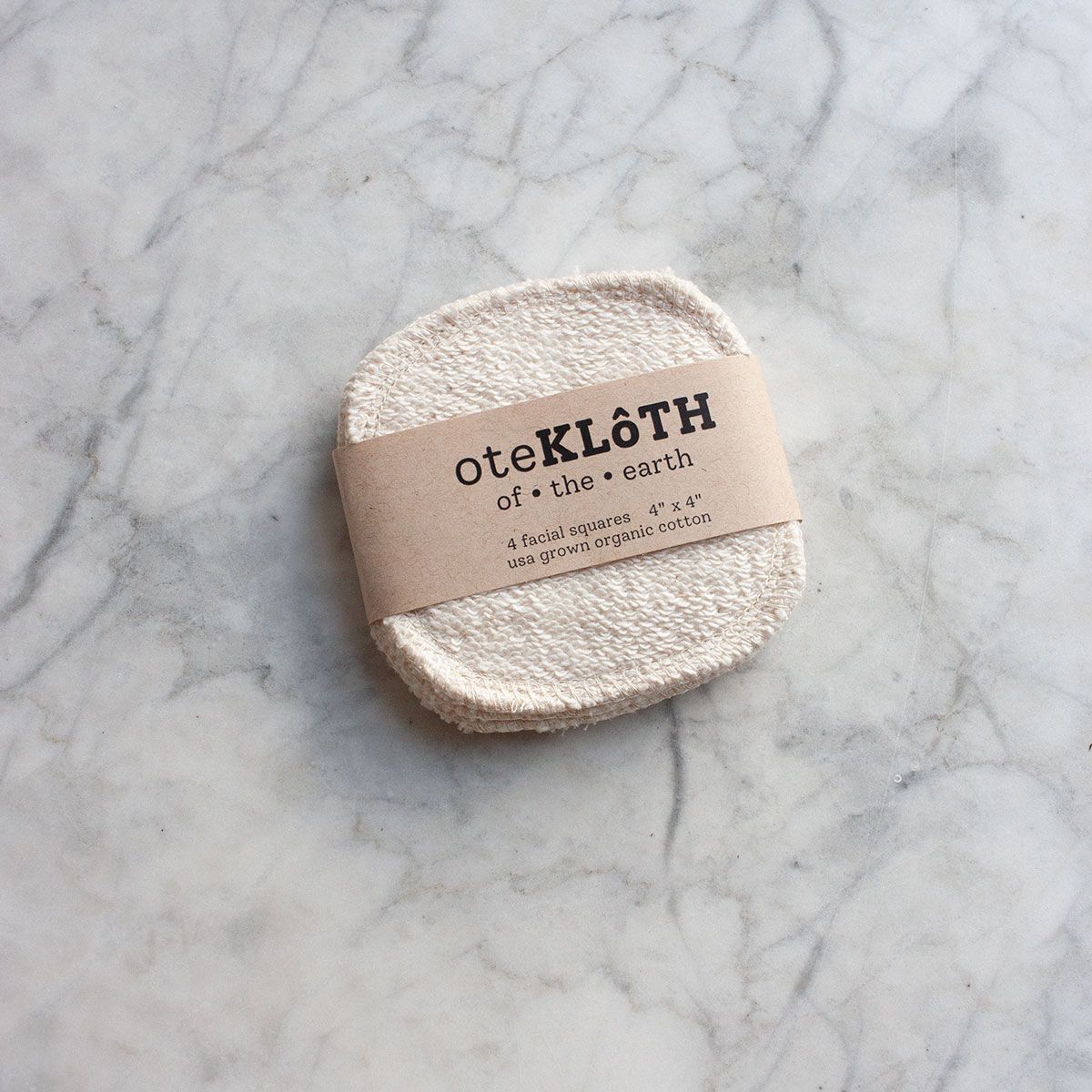 A set of woven, textured cotton squares are minimally packaged with a strip of brown paper reading: "oteKLôTH | of. the. earth | 4 facial squares 4" x 4" | usa grown organic cotton" are placed at a slight angle on a marble counter top.