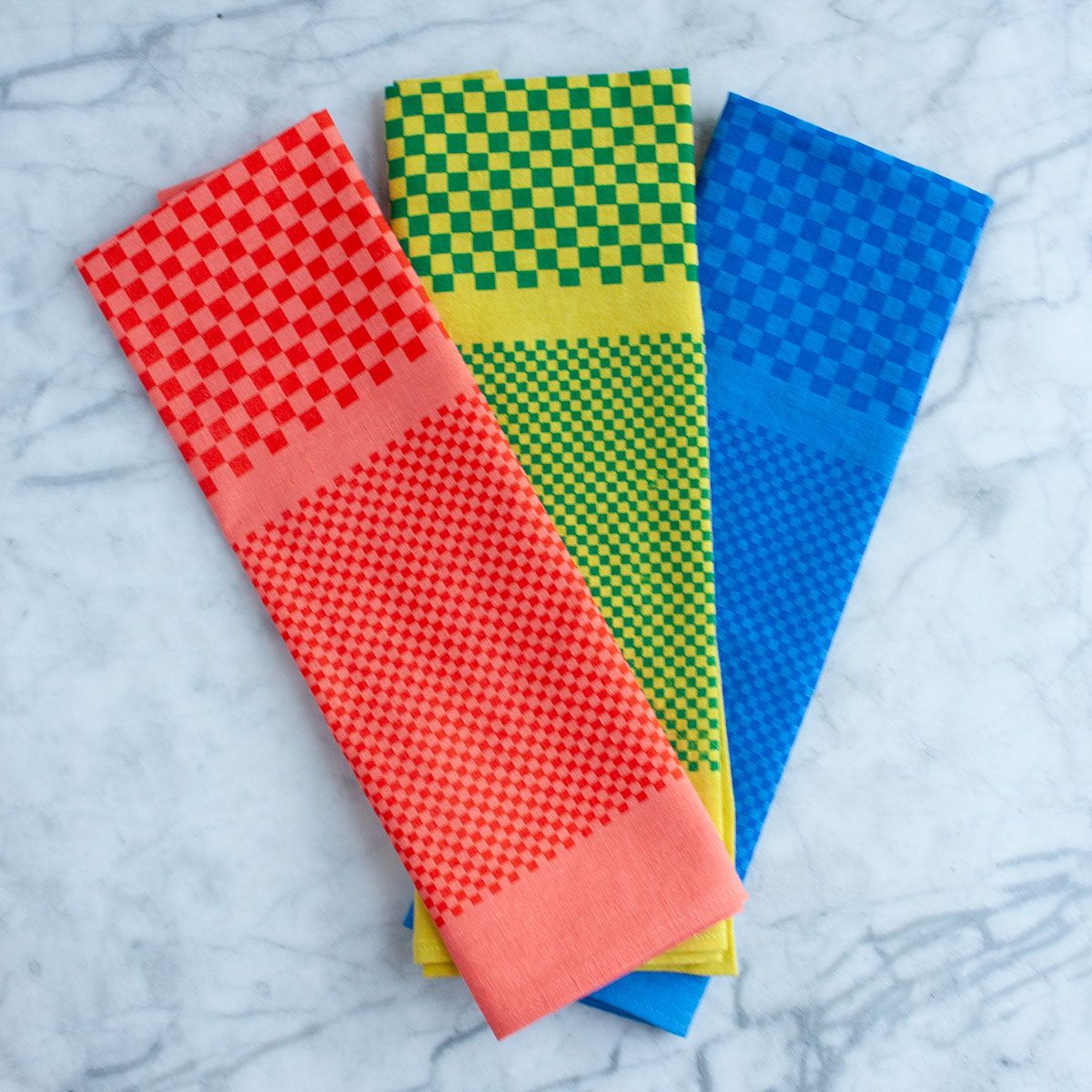 Three linen tea towels with monochromatic checker prints are fanned out and resting on a marble table top. The towels are shown, from left to right, in coral, green, and cobalt.