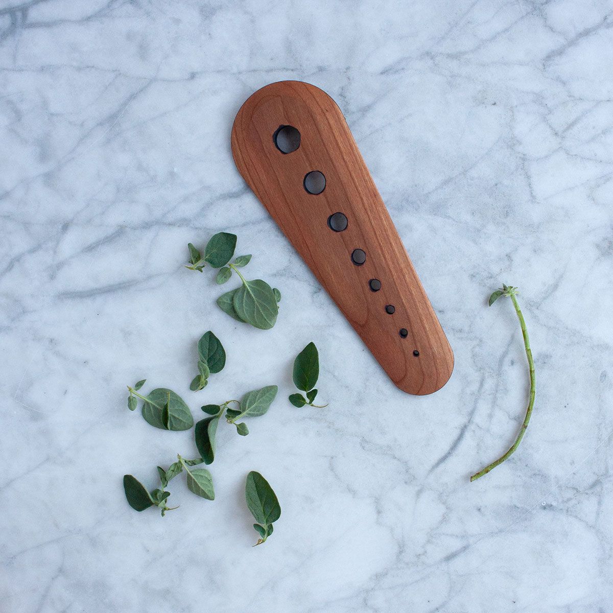 Cherry herb stripper sitting on a marble table top is surrounded by de-stemmed oregano and a leafless stem.