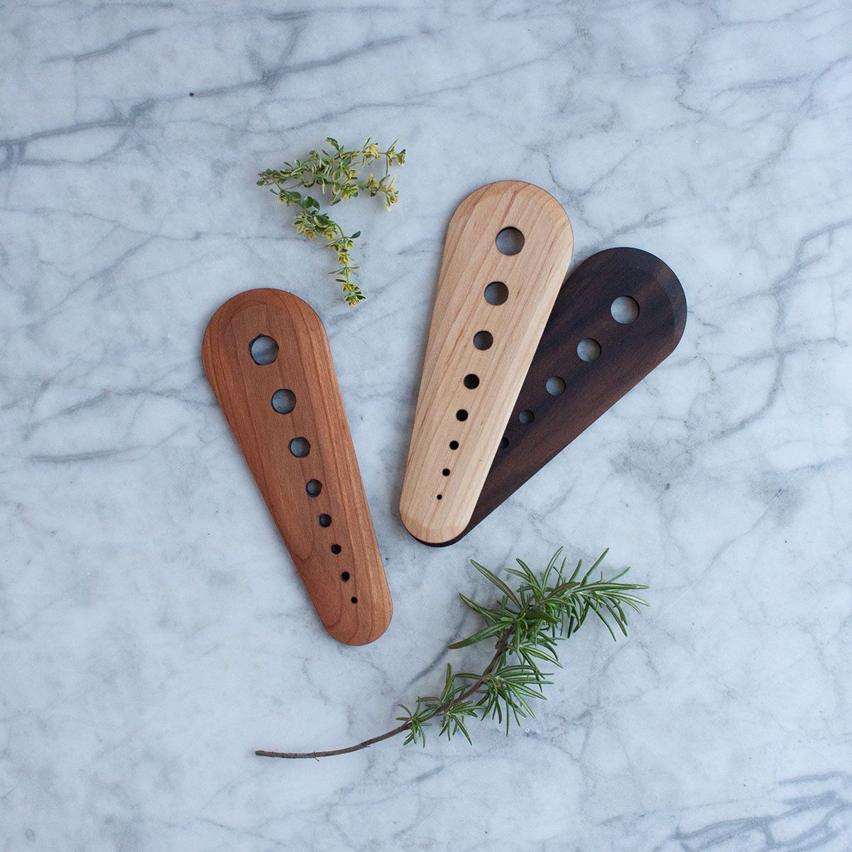 Three herb strippers, oblong solid wood pieces with 8 holes in gradually increasing circumference, in the available wood types: cherry, maple, and walnut rest on a marble slab with fresh herbs framing them.