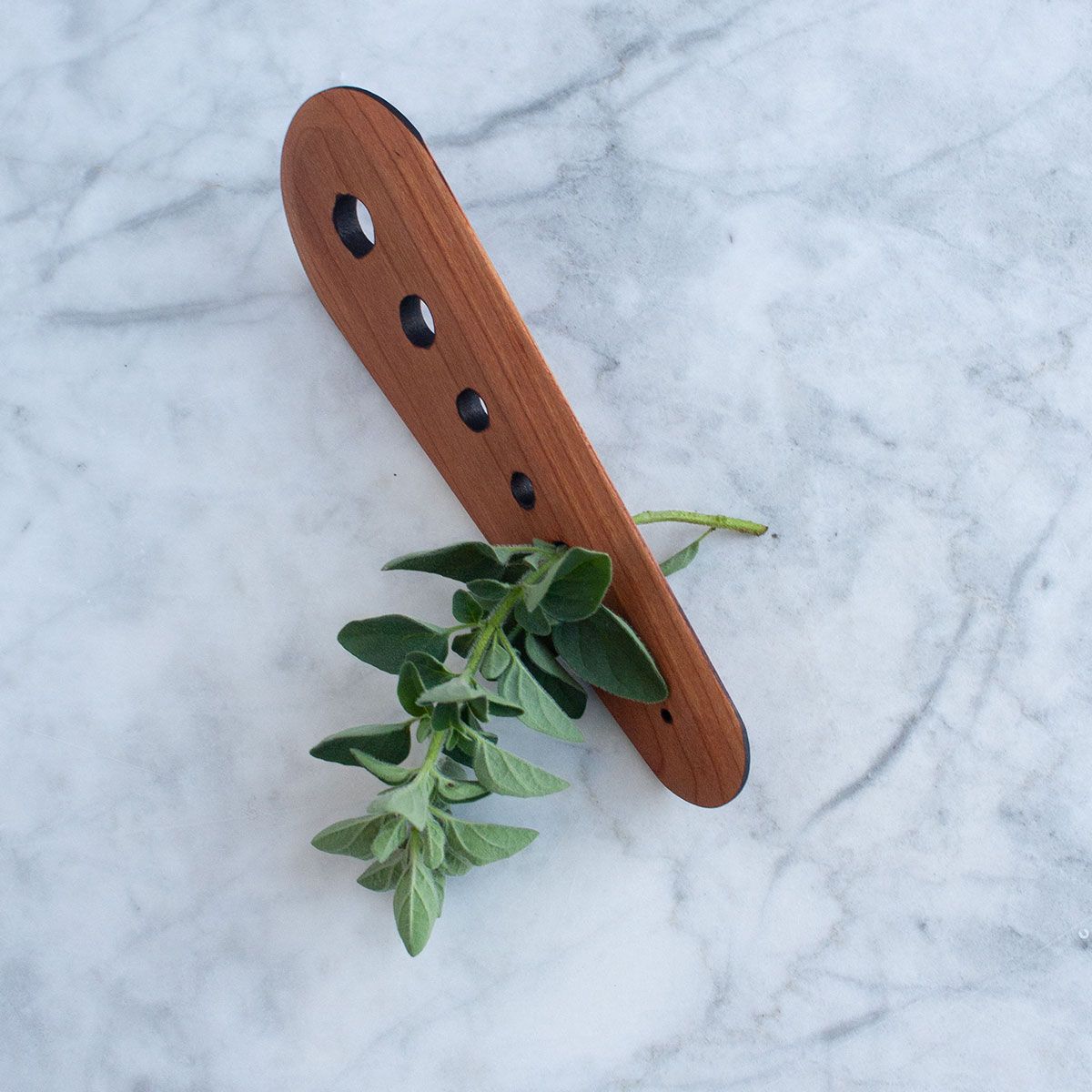 Herb stripper in cherry rests on a marble surface with a sprig of oregano threaded through the third smallest hole.