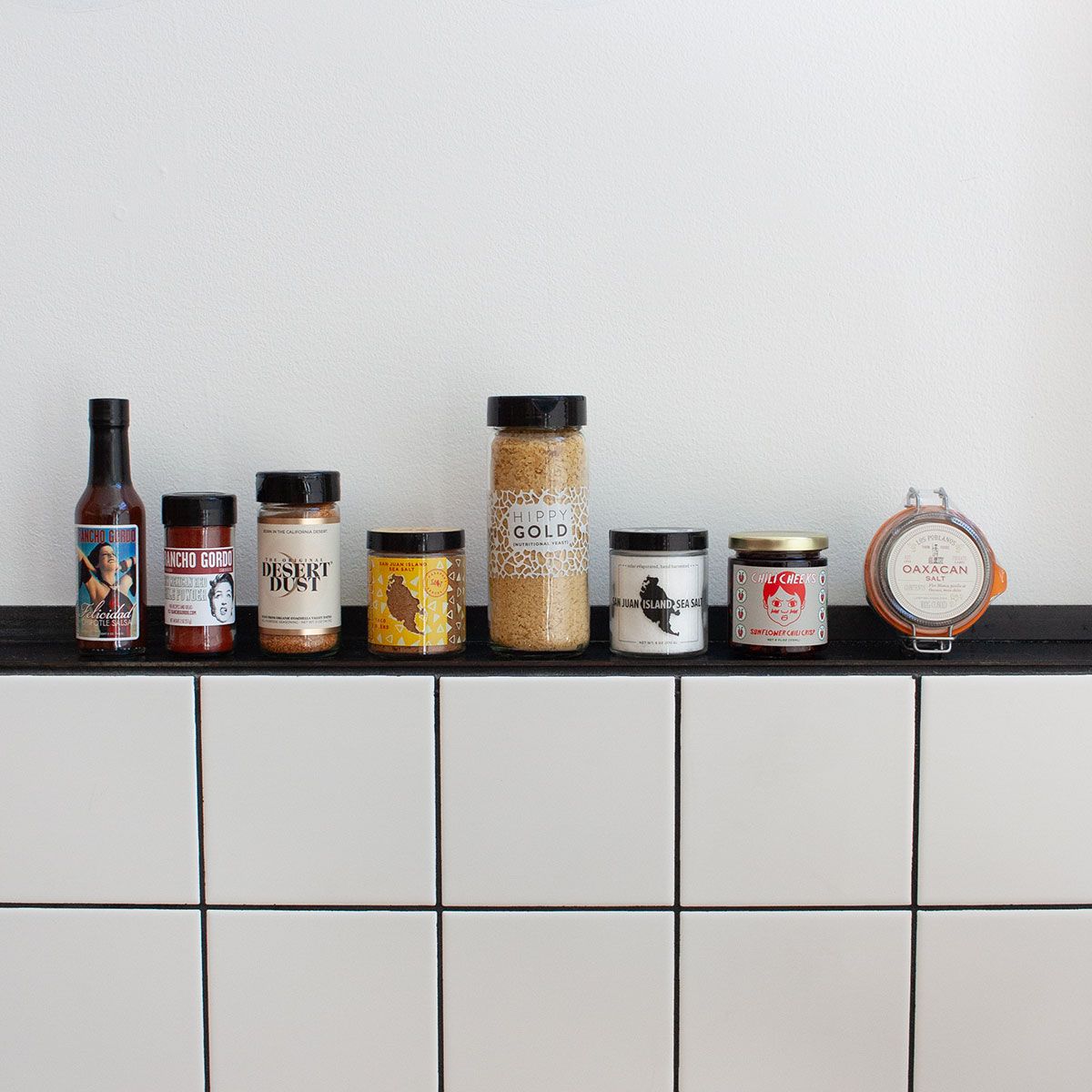 Sauces and seasonings pictured placed on a black metal ledge atop a wall of white tile with black grout and a white wall above. From left to right: Rancho Gordo Chipotle Salsa. Rancho Gordo Chili Powder, Desert Dust, San Juan Sea Island Sea Salt Taco Blend, San Juan Island Salt Hippy Gold Nutritional Yeast, San Juan Island Salt Evaporated Sea Salt, Chili Cheeks, and Los Poblanos Oaxaca Salt