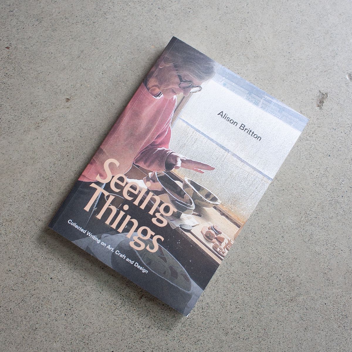 Book titled "Seeing Things" by Alison Britton placed on a grey textured surface. The cover features a photograph of an older woman in a pink sweater and glasses, working with ceramic pieces. The subtitle reads "Collected Writing on Art, Craft and Design."