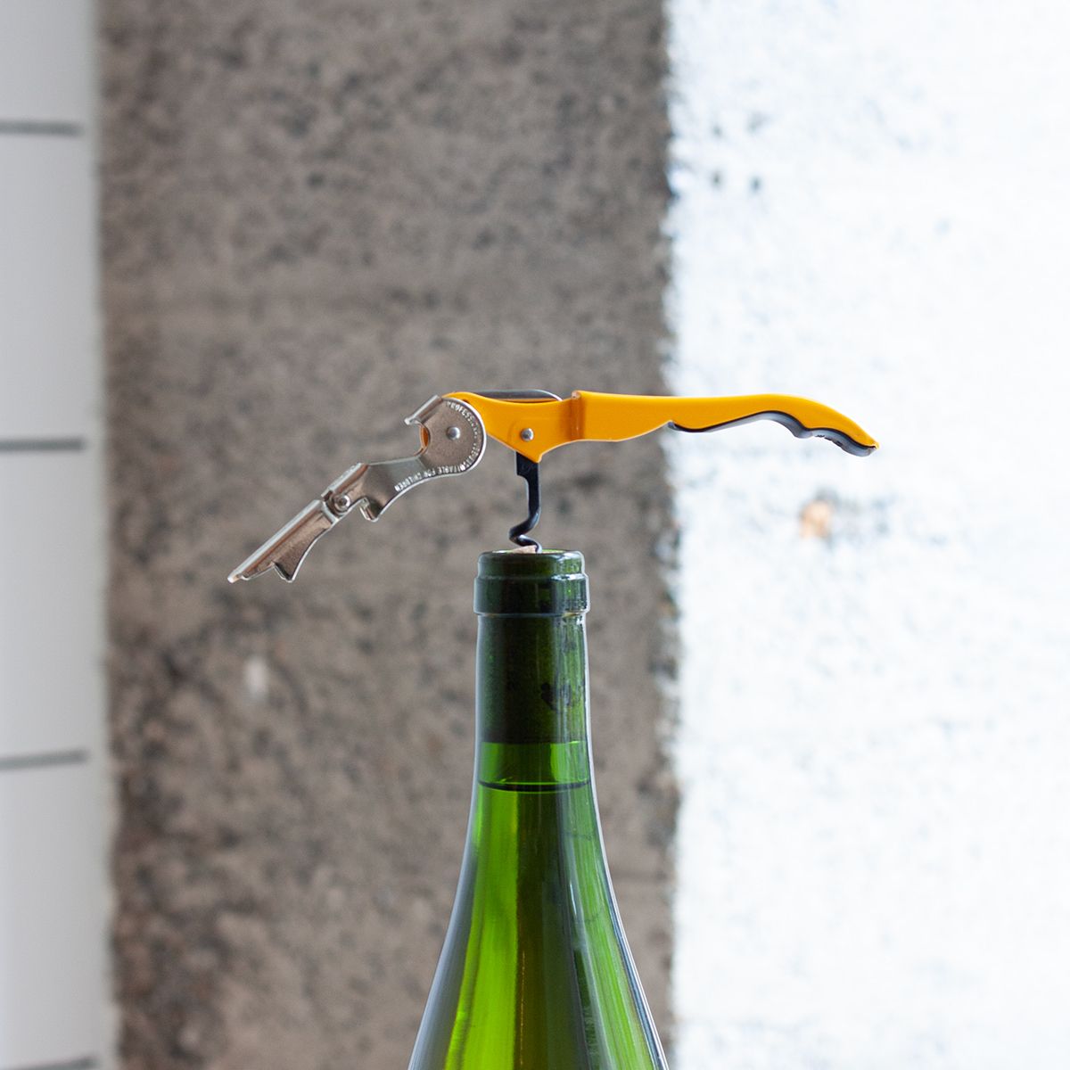 A yellow corkscrew is in action- screwed into the cork of a green glass wine bottle against a gray concrete background.