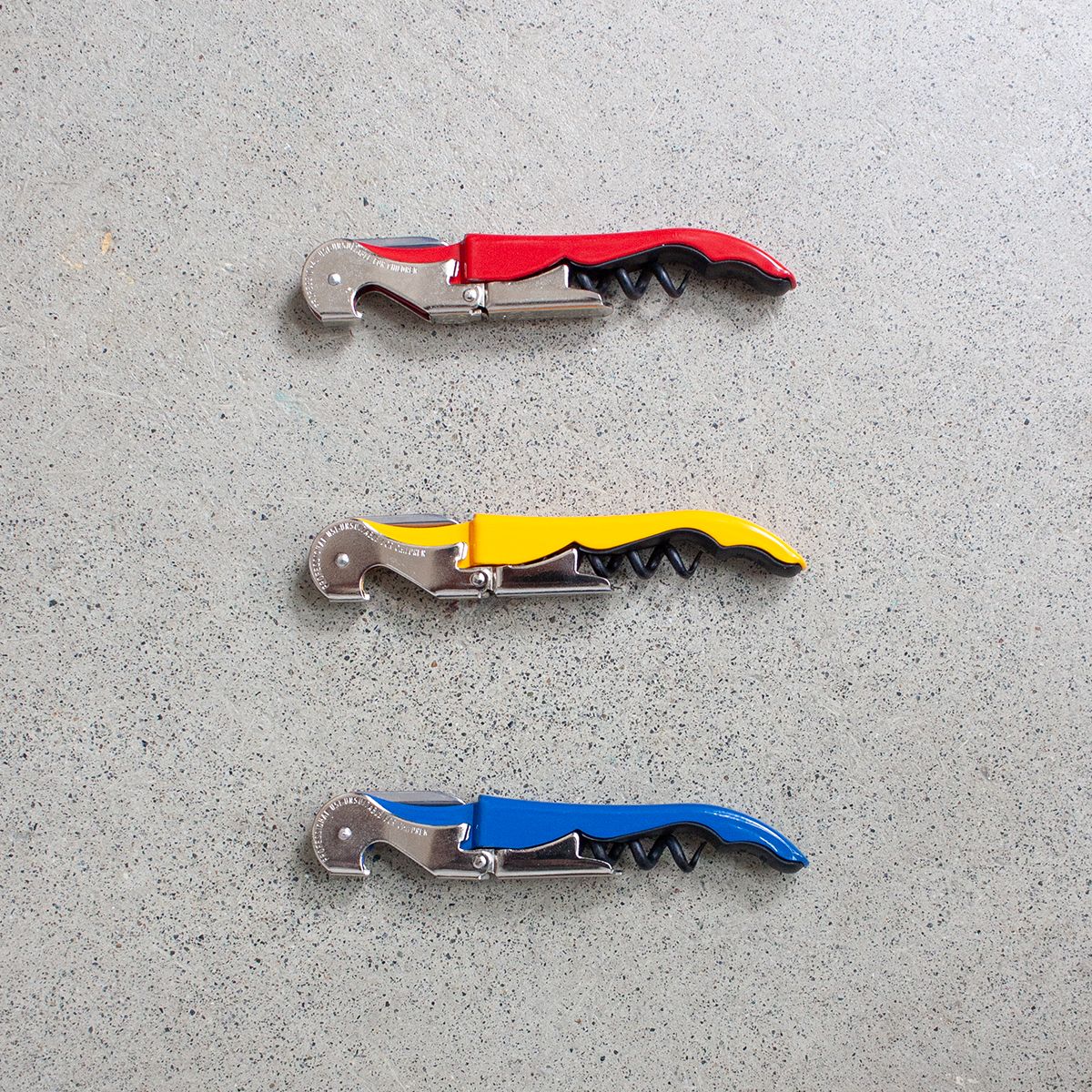 A red, yellow, and blue corkscrew are placed parallel to each other on a concrete surface.