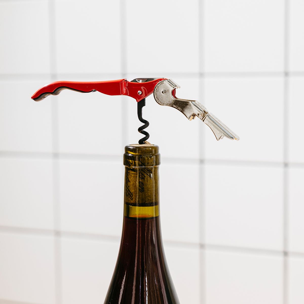 A red corkscrew is in action- screwed into the cork of a green glass wine bottle against a white tile background.