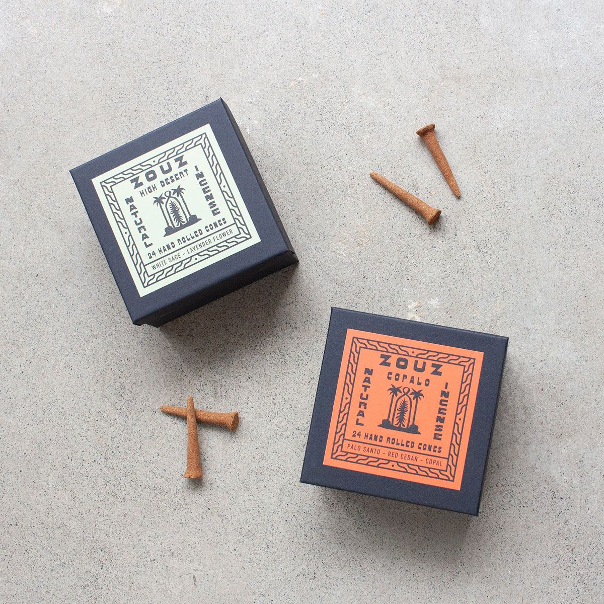 Two black boxes of Zouz incense on a concrete surface, one labeled "High Desert" with white sage and lavender, the other labeled "Copalo" with palo santo and red cedar. Four incense cones are placed around the boxes.
