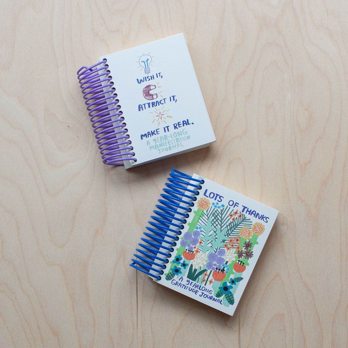 Two spiral-bound journals on a wooden surface: one with a purple coil titled "Wish it, Attract it, Make it Real," and another with a blue coil titled "Lots of Thanks" featuring floral illustrations.