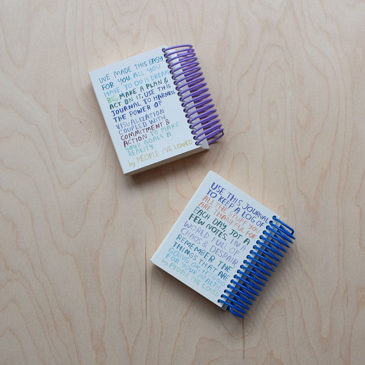 Two spiral-bound journals on a wooden surface: one with a purple coil and another with a blue coil with colorful text filling the back page.