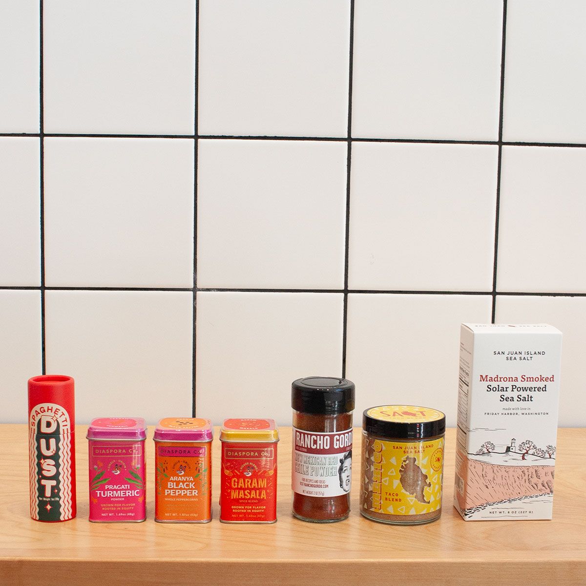 Various spices and salts arranged in a straight line on a light wood surface against a white tile background.