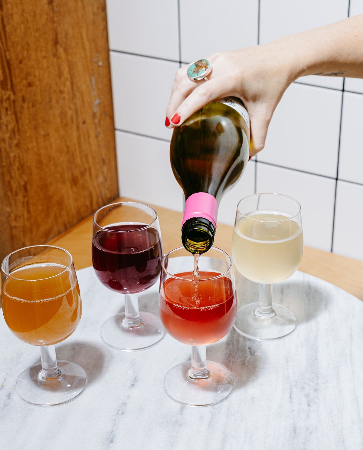 A hand pours wine into a half-filled glass sitting on a marble table top with other glasses of wine on the table as well. There is a wood beam and white tile wall in the background.