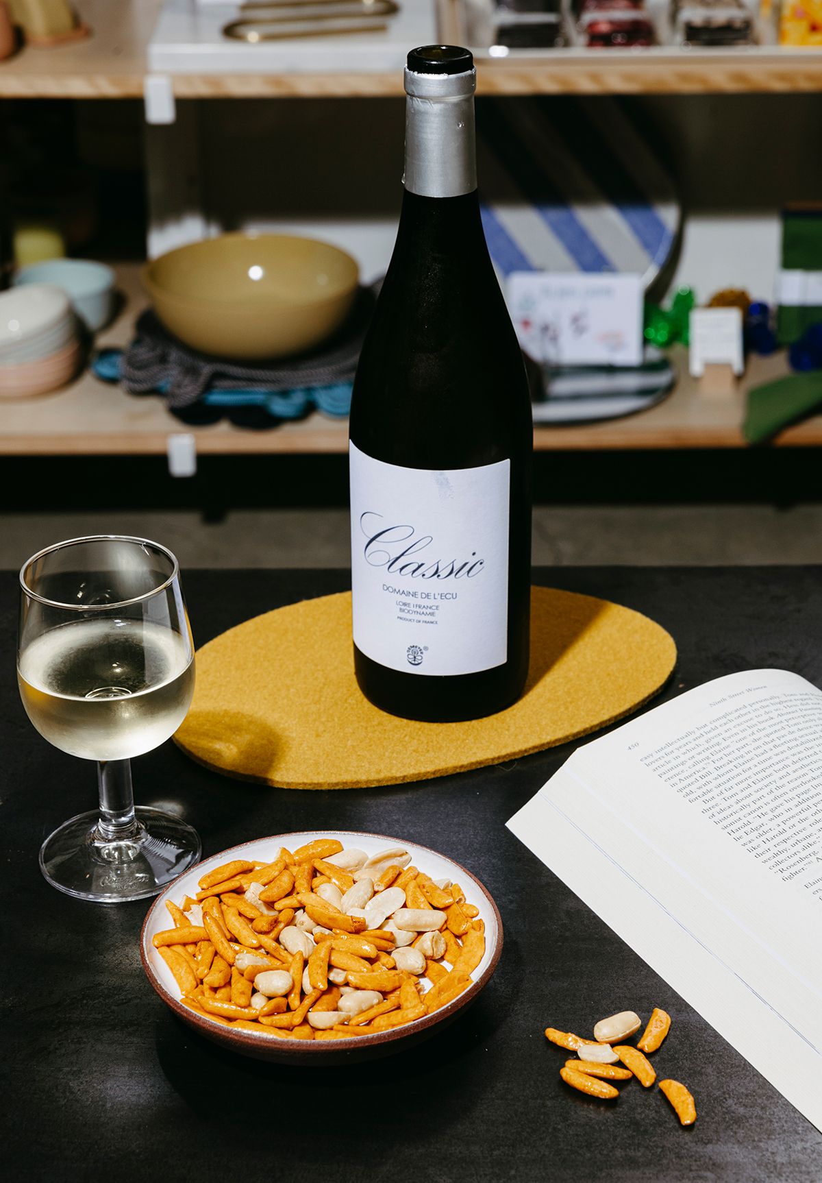 A bottle of wine with a white label sits upon a yellow felted ovular trivet on a metal table top. Nearby sits a glass of wine, a book, and a dish filled with a snack mix.