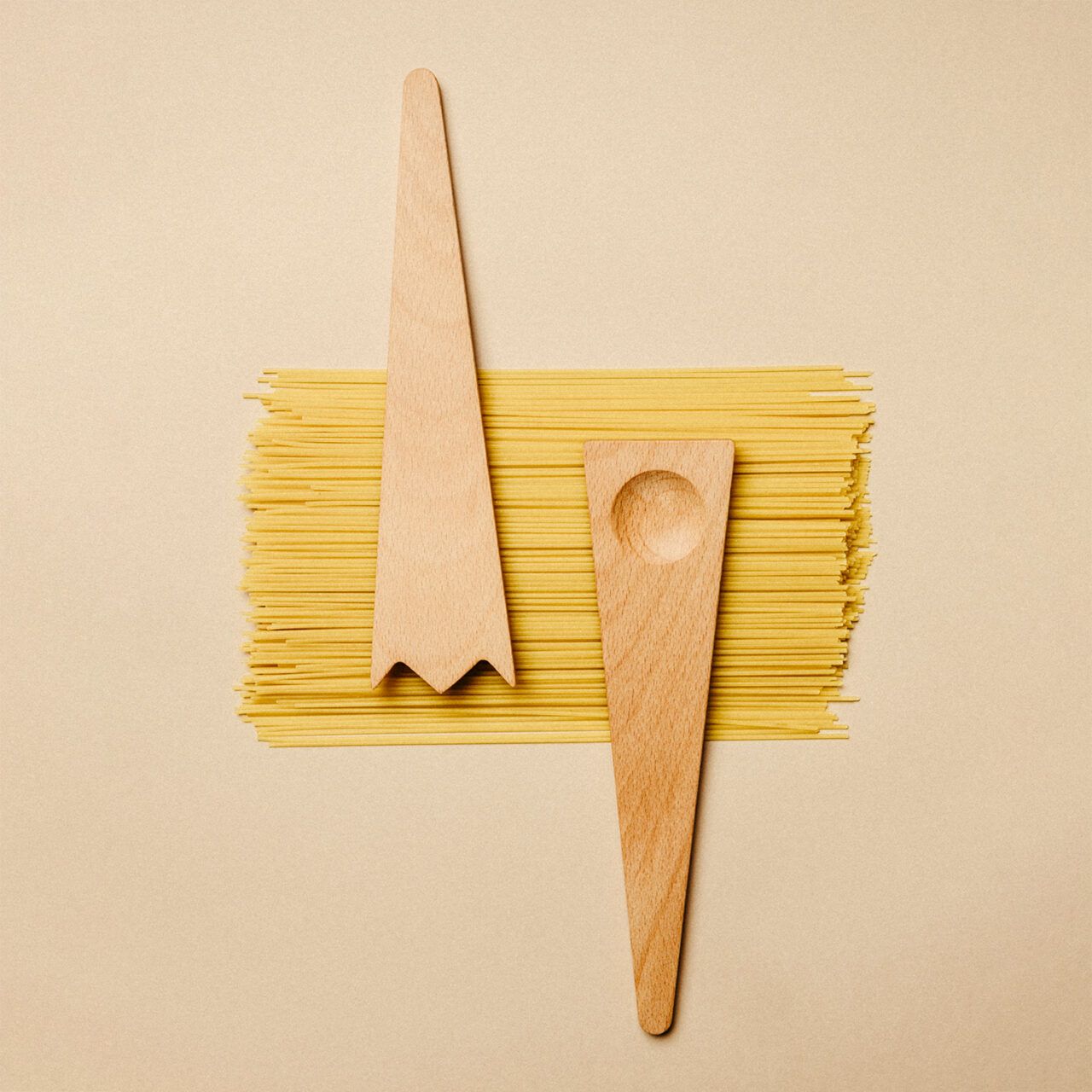 Two wooden salad servers placed on a bundle of uncooked spaghetti noodles on a beige background.