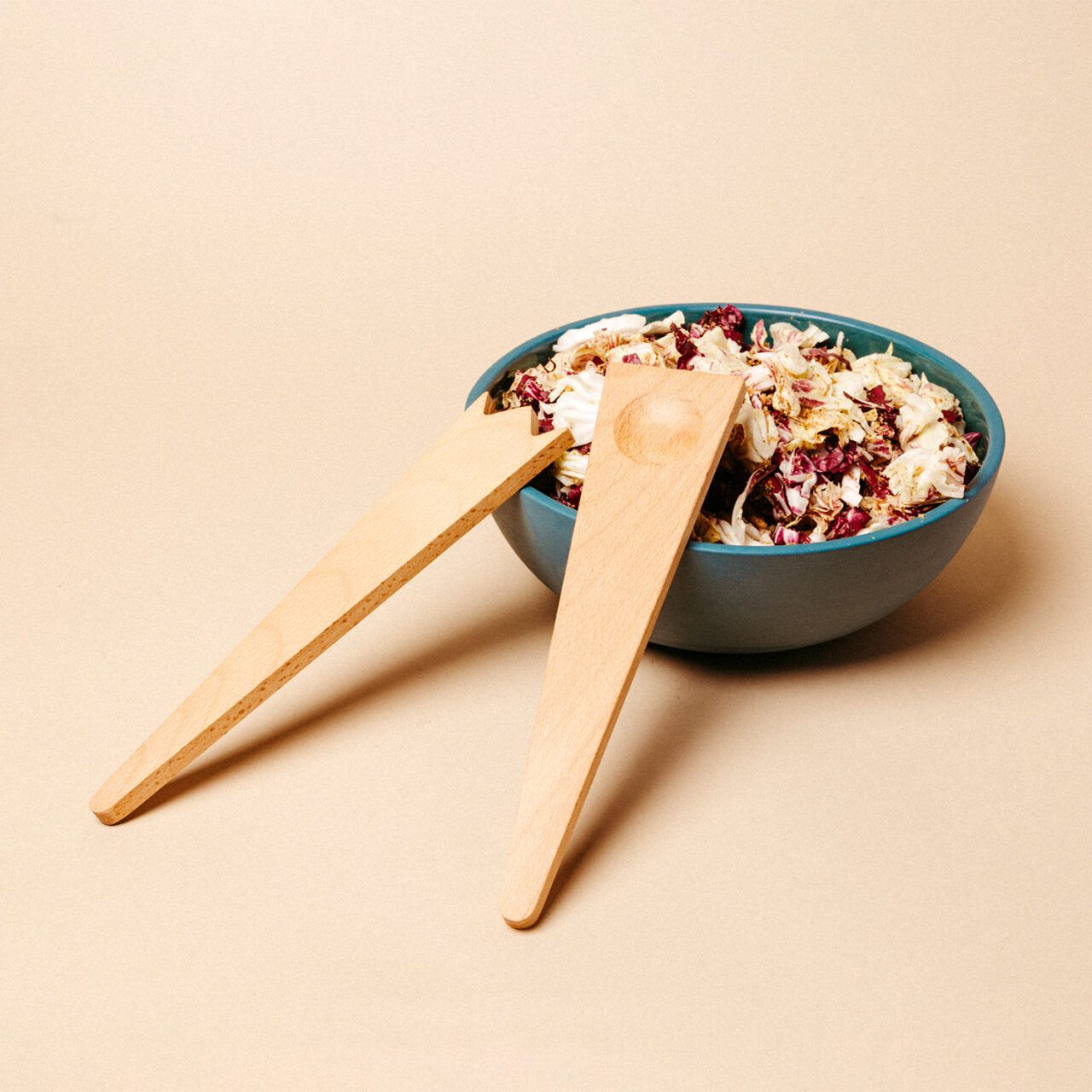 Two wooden salad servers placed on the edge of a large blue bowl holding salad, on a beige background.