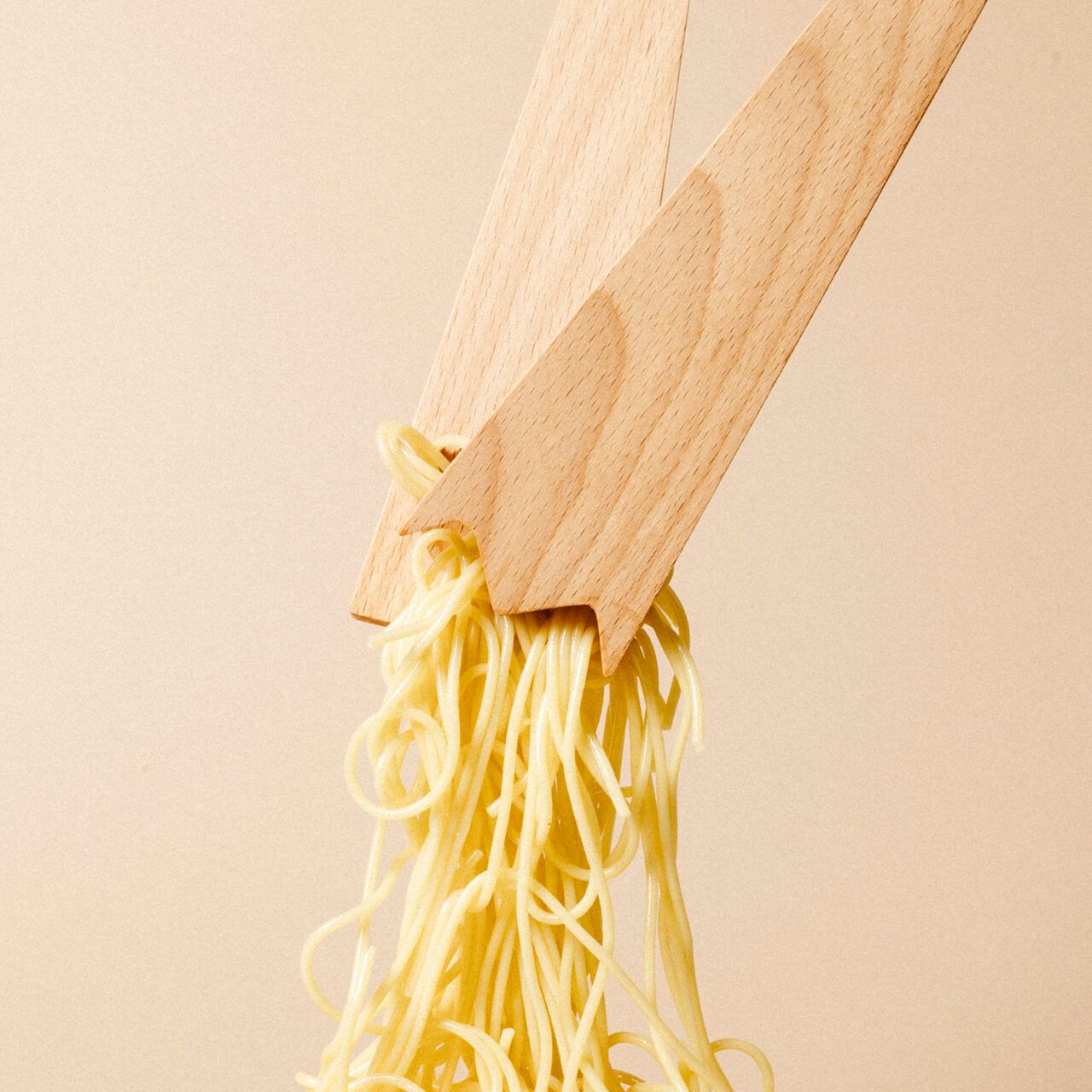 A pair of wooden salad servers lifting cooked spaghetti against a beige background.