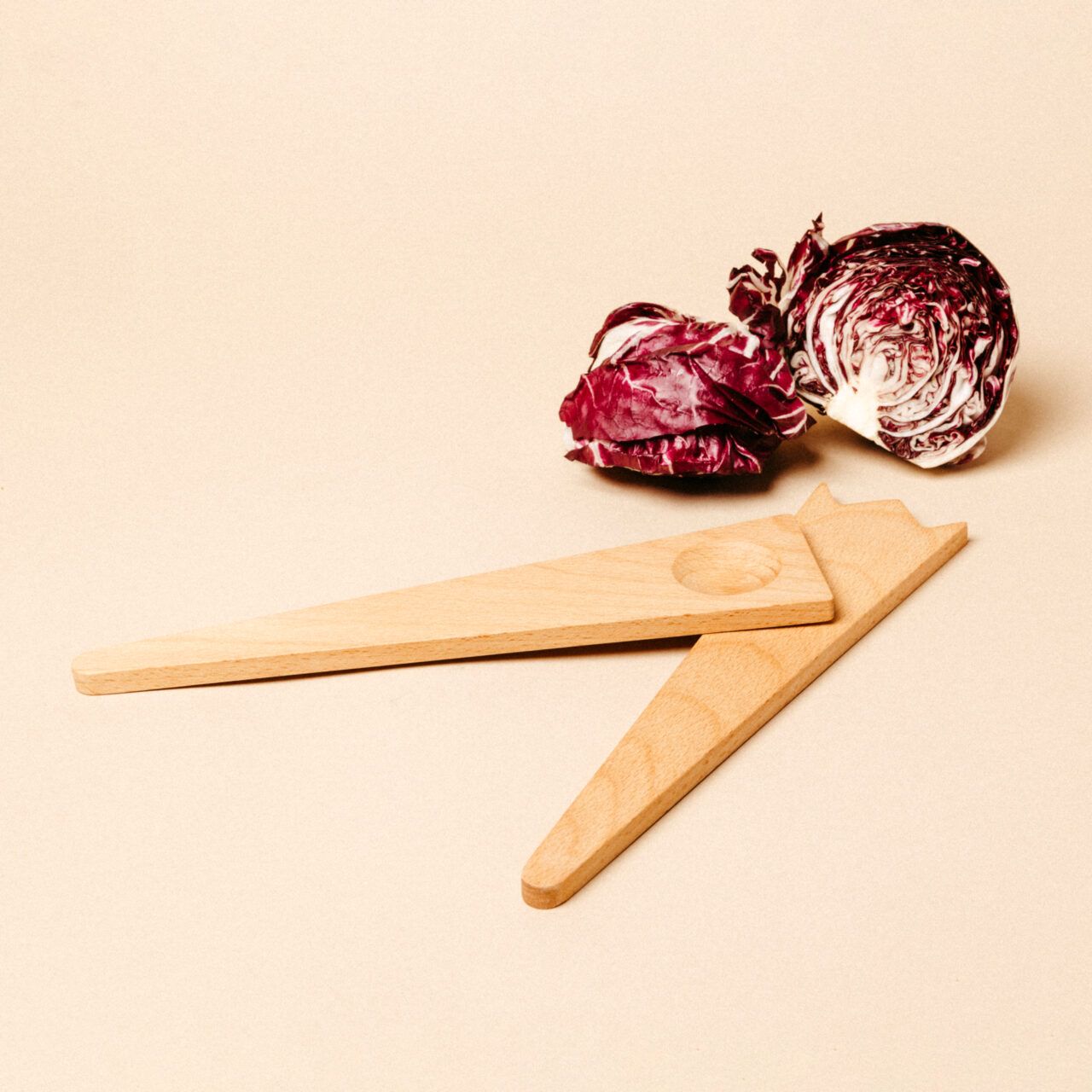 Two wooden salad servers placed near a head of red radicchio that has been sliced into two pieces on a beige background.