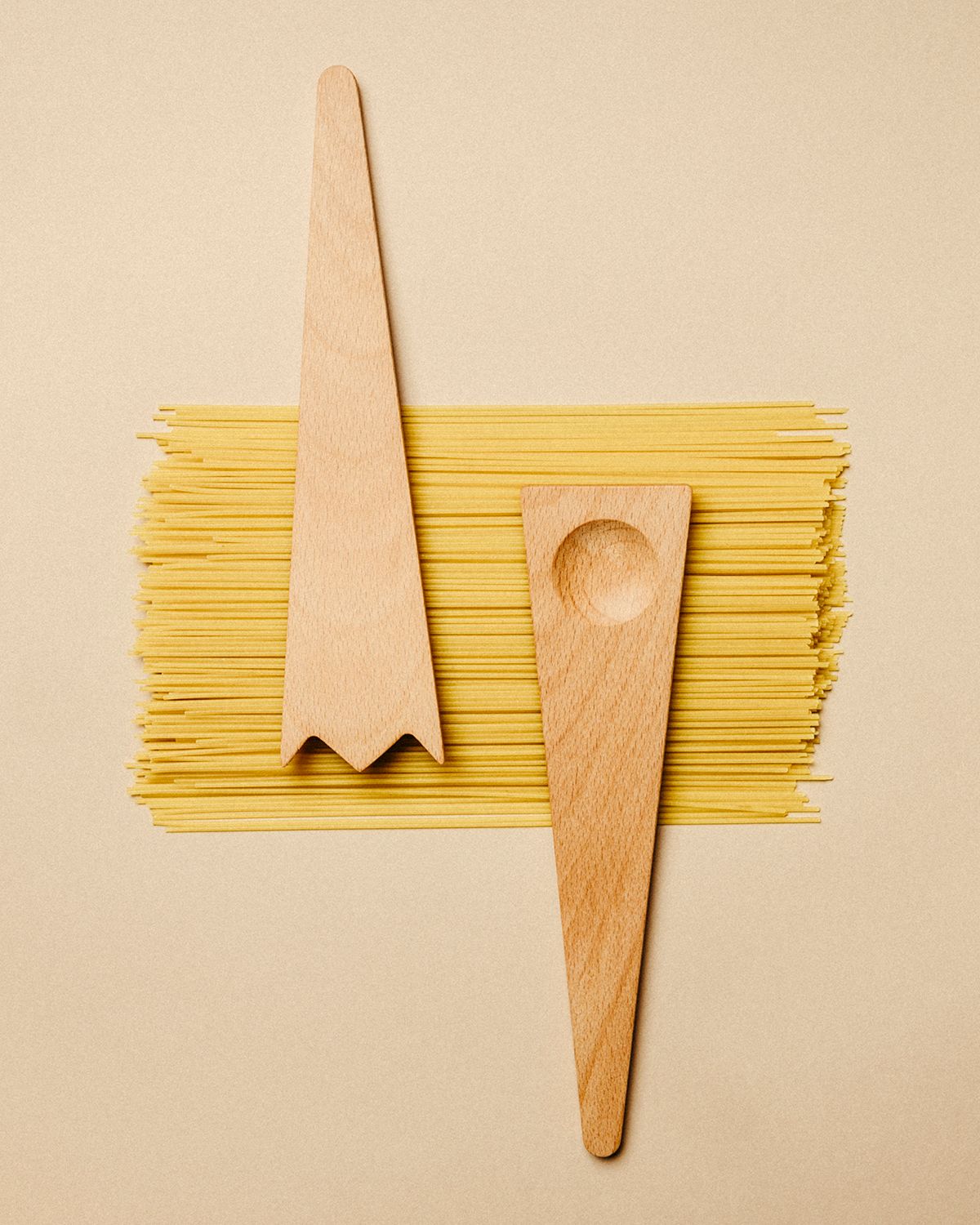 Wooden salad servers lying on a stack of uncooked spaghetti against a neutral backdrop.