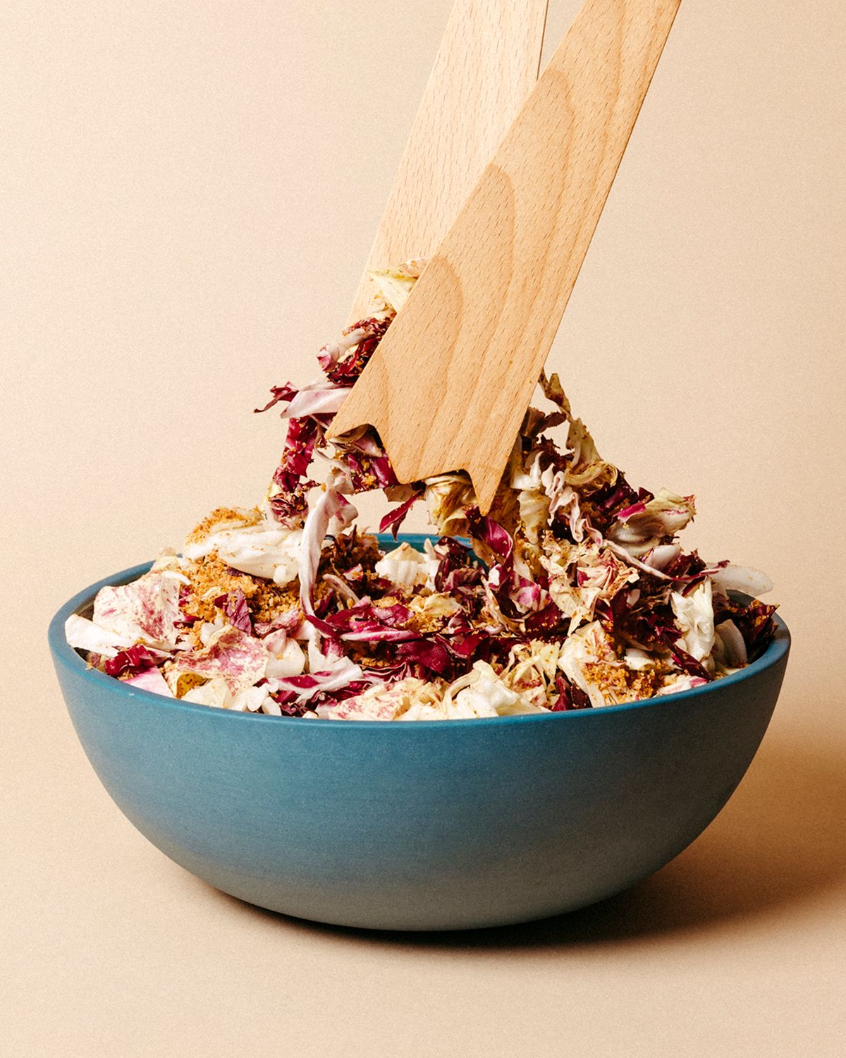 Wooden salad servers in action, tossing a radicchio salad in a blue bowl on a neutral backdrop.