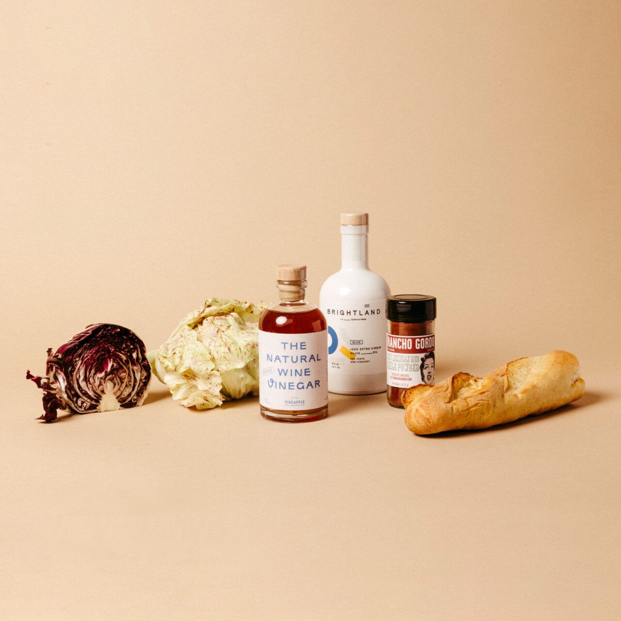 An assortment of food items displayed on a beige background: red cabbage, green and red speckled radicchio, natural wine vinegar, extra virgin olive oil, seasoning, and a loaf of bread.