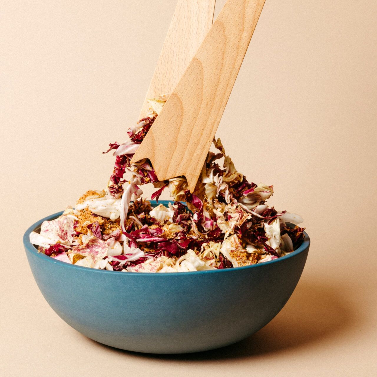 Wooden salad servers in action, tossing a radicchio salad in a blue bowl on a neutral backdrop.