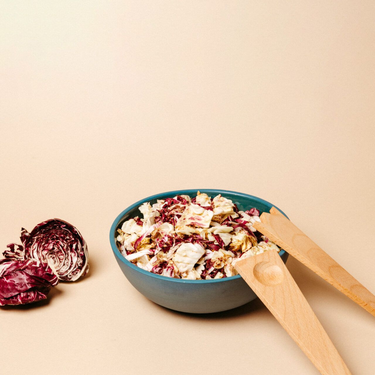 Two wooden salad servers placed on the edge of a large blue bowl holding salad, on a beige background with a sliced red radicchio nearby.