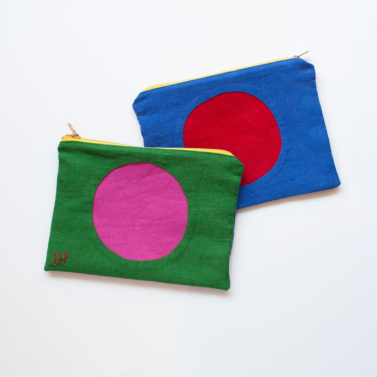 Two colorful fabric pouches with zipper closures, arranged on a white background. The top pouch shows the blue with a large red circle in the center side, while the bottom pouch displays the side of the pouch that is green with a large pink circle. Featuring yellow zippers, and the green side has the initials "DF" embroidered in red on the bottom left corner.