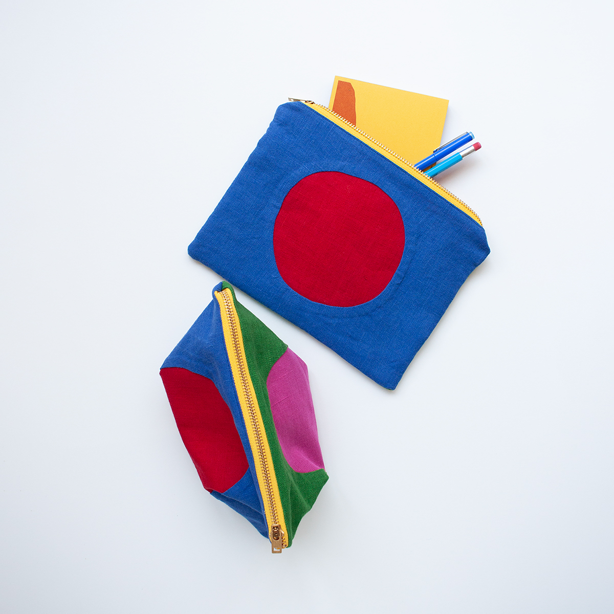 Two colorful fabric pouches with yellow zippers, arranged on a white background. The top pouch lays flat and is blue with a red circle in the center and has a yellow card and two pens partially visible inside. The bottom pouch is displayed from a different angle, showing its combination of blue, green, and red panels with a pink circle.