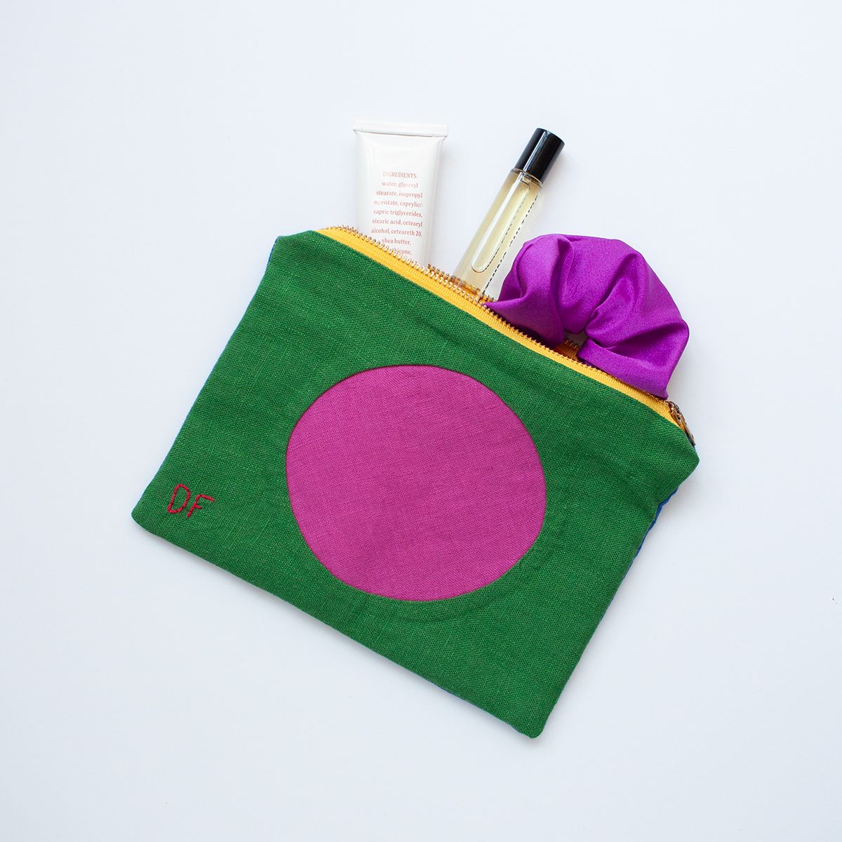 A colorful fabric pouch a with a yellow zipper closure is arranged on a white background. The pouch shows green fabric with a large pink circle with the initials "DF" in red stitching on the bottom left corner. The bag is open and a purple scrunchie, small perfume bottle, and tube of hand cream are partially showing out of the bag.